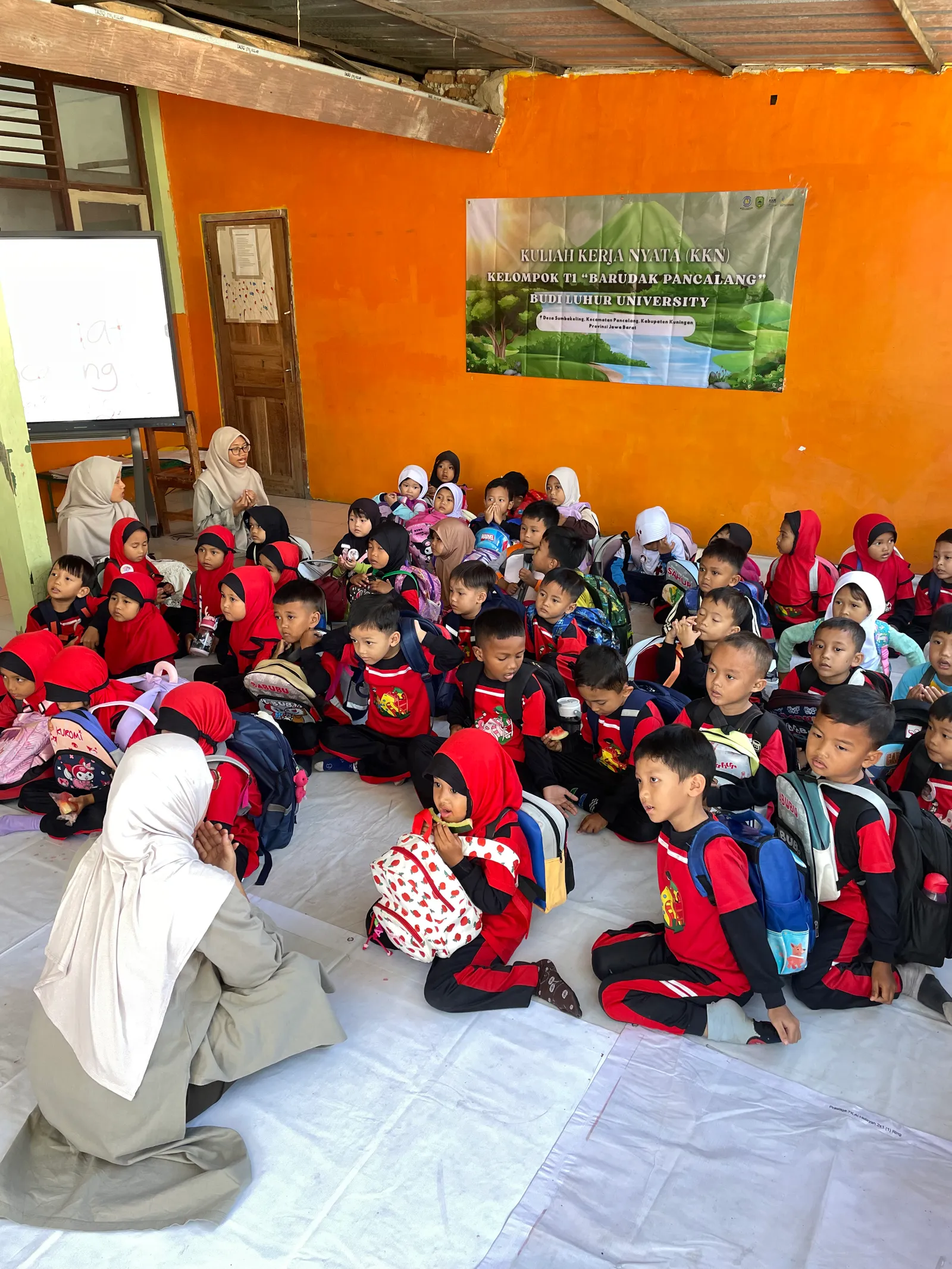 Peserta didik TK Al-Hidayah duduk bersama sambil membawa tas belajar yang dibagikan tim KKN T1.