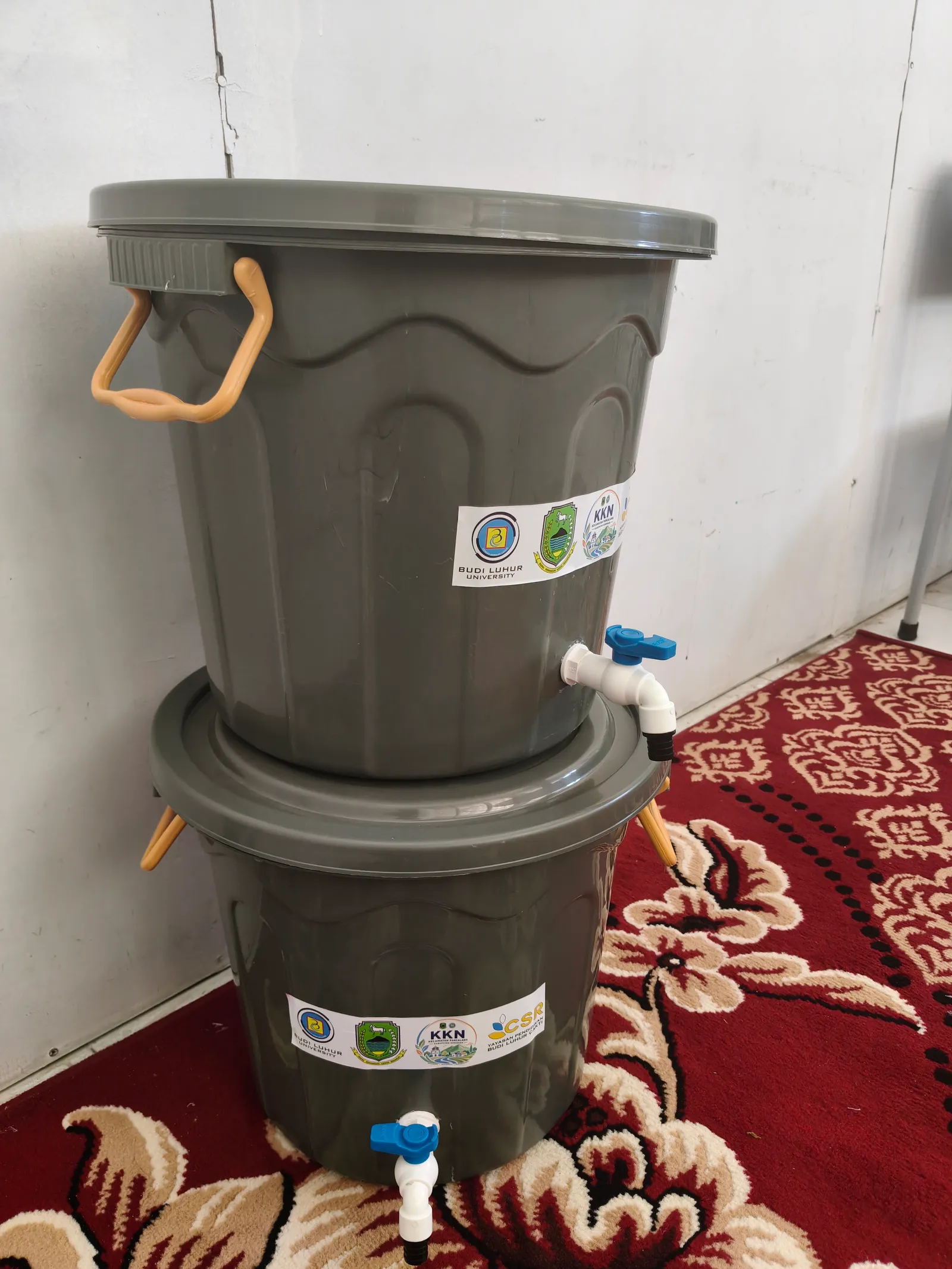 Dua tong keran air berwarna abu-abu dengan label KKN dan CSR untuk TK Al-Hidayah.